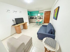 A living room with a sofa, a TV on a stand, a small table, and a kitchen in the background. - Marupiara Muro Alto (Ipojuca)