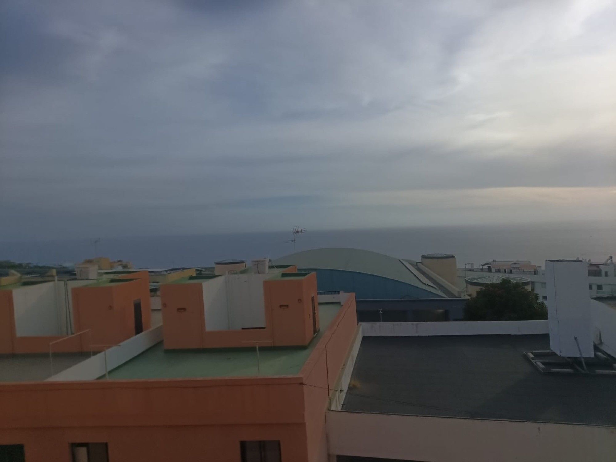 Una azotea con edificios al fondo, cielo despejado y vista distante del mar.