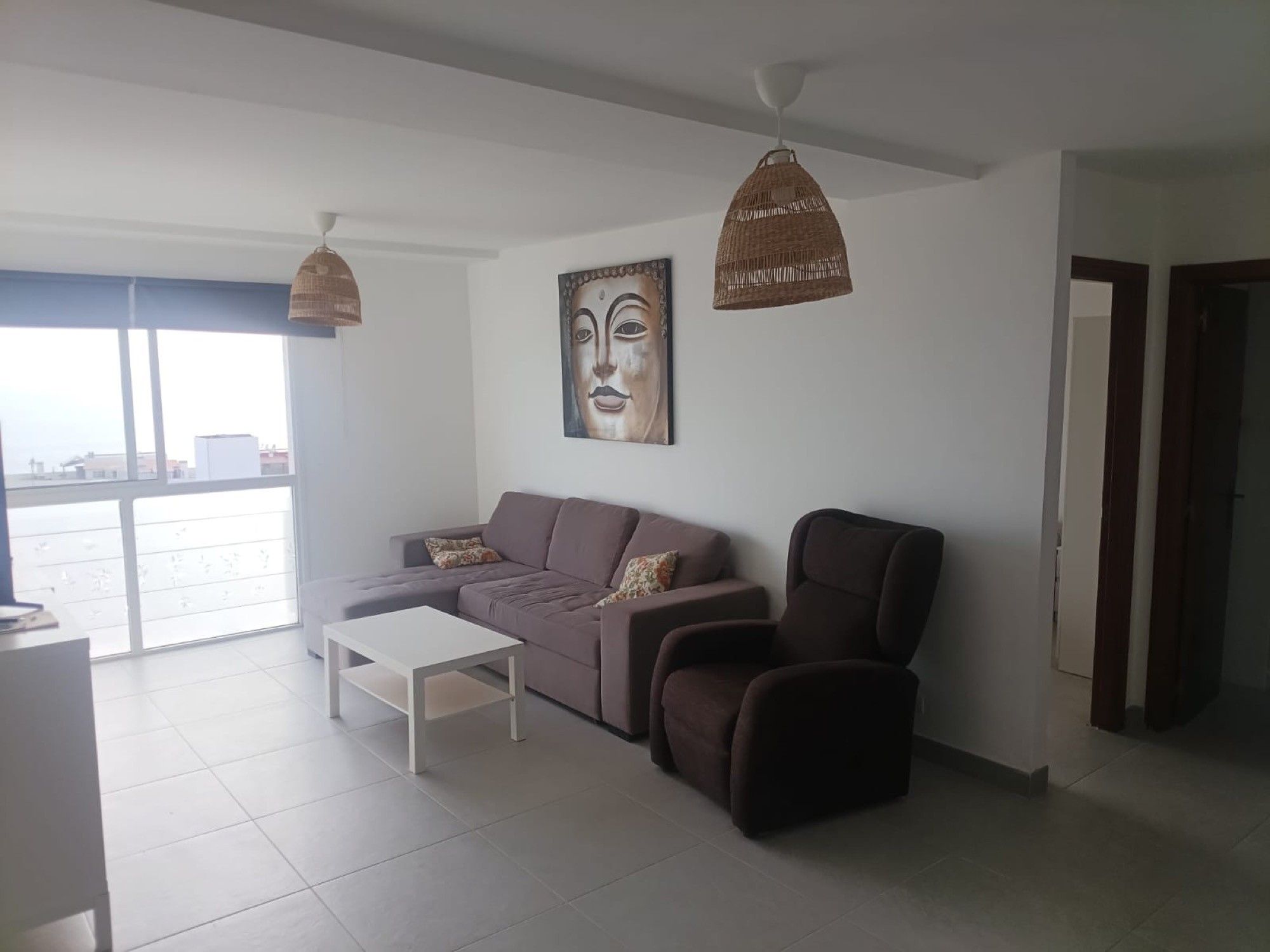 Una sala de estar moderna con sofá, sillón y mesa de centro. Hay un ventanal grande que ofrece luz natural y vistas a la ciudad.
