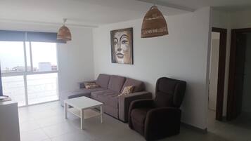 Una sala de estar moderna con sofá, sillón y mesa de centro. Hay un ventanal grande que ofrece luz natural y vistas a la ciudad.
