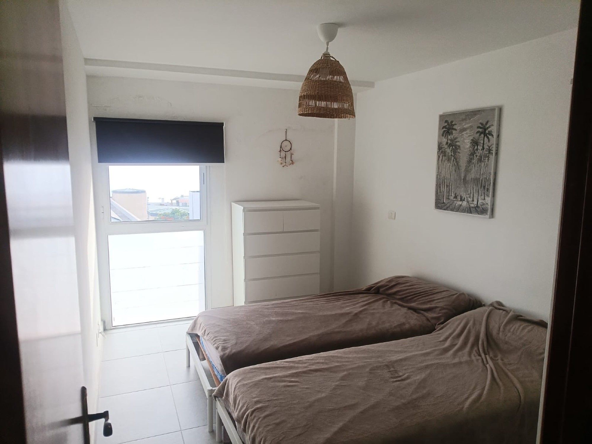 Un dormitorio con dos camas, una ventana con persianas, un aparador blanco, un atrapasueños colgante y un cuadro en la pared.