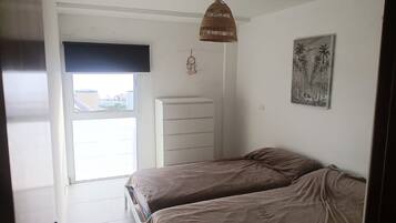 Un dormitorio con dos camas, una ventana con persianas, un aparador blanco, un atrapasueños colgante y un cuadro en la pared.