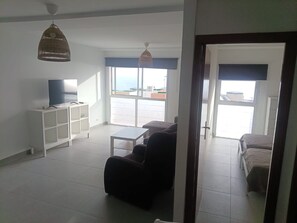 A living room with a TV, a sofa, a coffee table, and a bookshelf. There is a large window with a view of the outside.