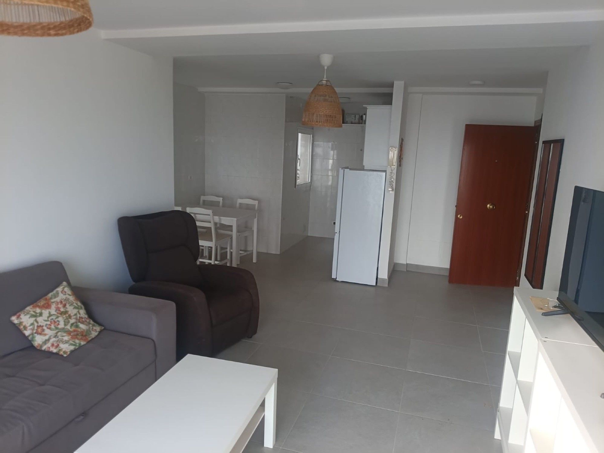 Una sala de estar con un sofá y una mesa de centro, un comedor con sillas, una cocina con un refrigerador y una puerta de madera.