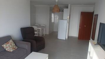 Una sala de estar con un sofá y una mesa de centro, un comedor con sillas, una cocina con un refrigerador y una puerta de madera.