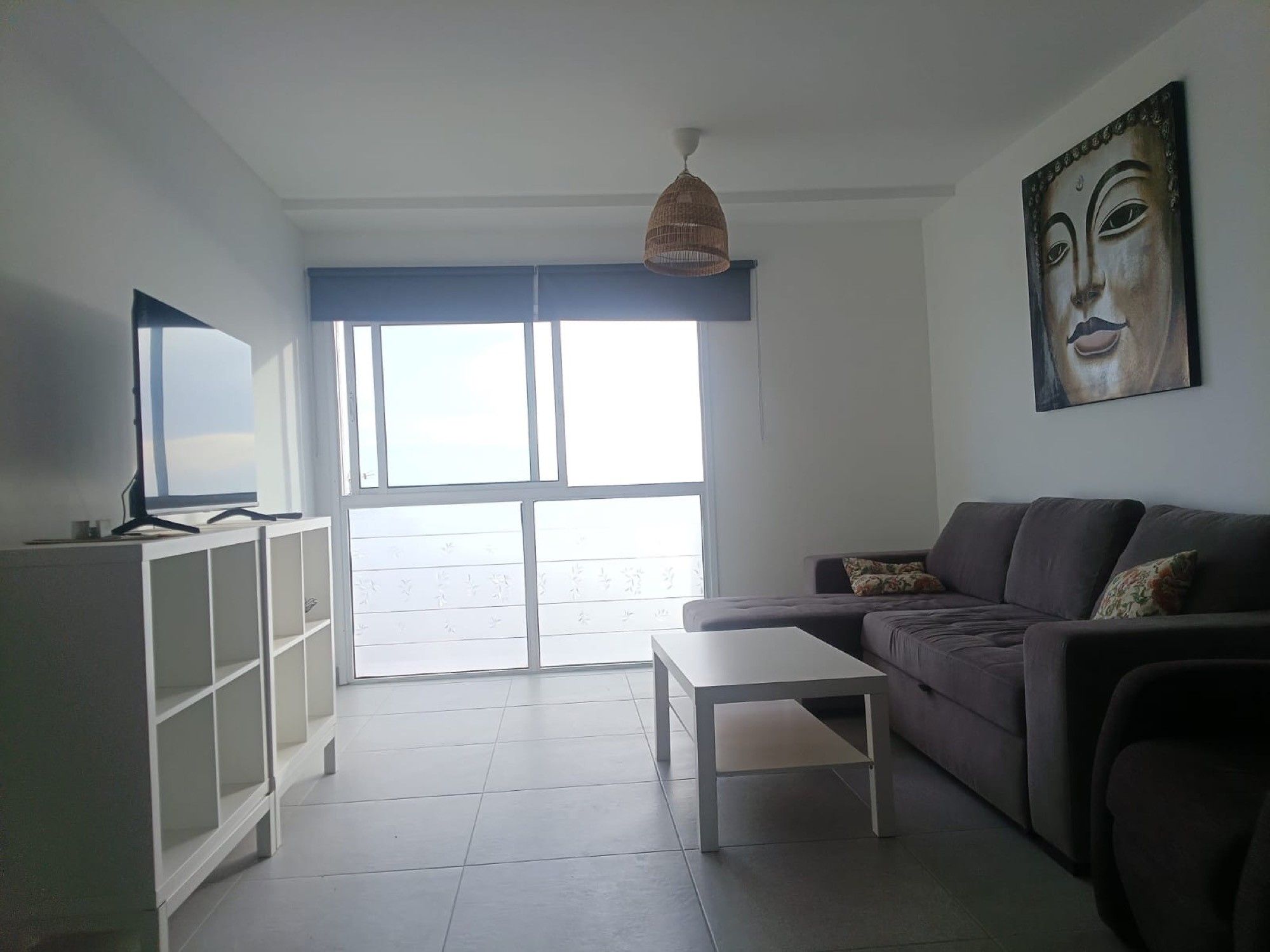 Una sala de estar con un sofá, una mesa de centro, un televisor sobre un mueble bajo y un ventanal grande.