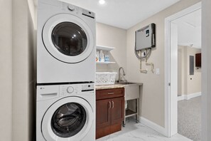 A laundry room with two stacked front-loading washing machines, a small sink with a faucet, a white laundry basket, and a wall-mounted heater. - Spectacular renovated 3-bedroom house in Jax Beach with pool patio & yar (Jacksonville Beach)