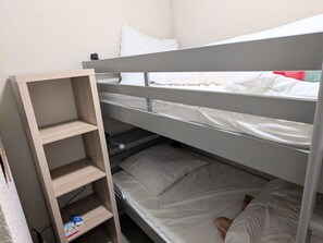 Ein Etagenbett mit einem Holzregal daneben, ein Einzelbett darunter und ein an der Wand befestigtes Regal.