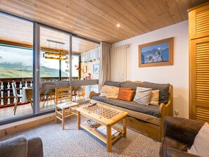 Ein gemütliches Wohnzimmer mit Holzbalkendecke, einer Couch mit Kissen, einem Couchtisch und Blick auf eine Berglandschaft durch große Fenster.