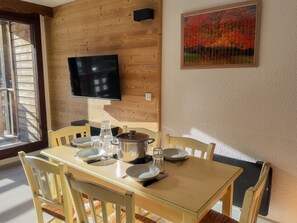 A dining area with a wooden table set for four, a television mounted on the wall, and a painting of autumn trees. - Bright apartment near slopes - 6 guests (Villarodin-Bourget)