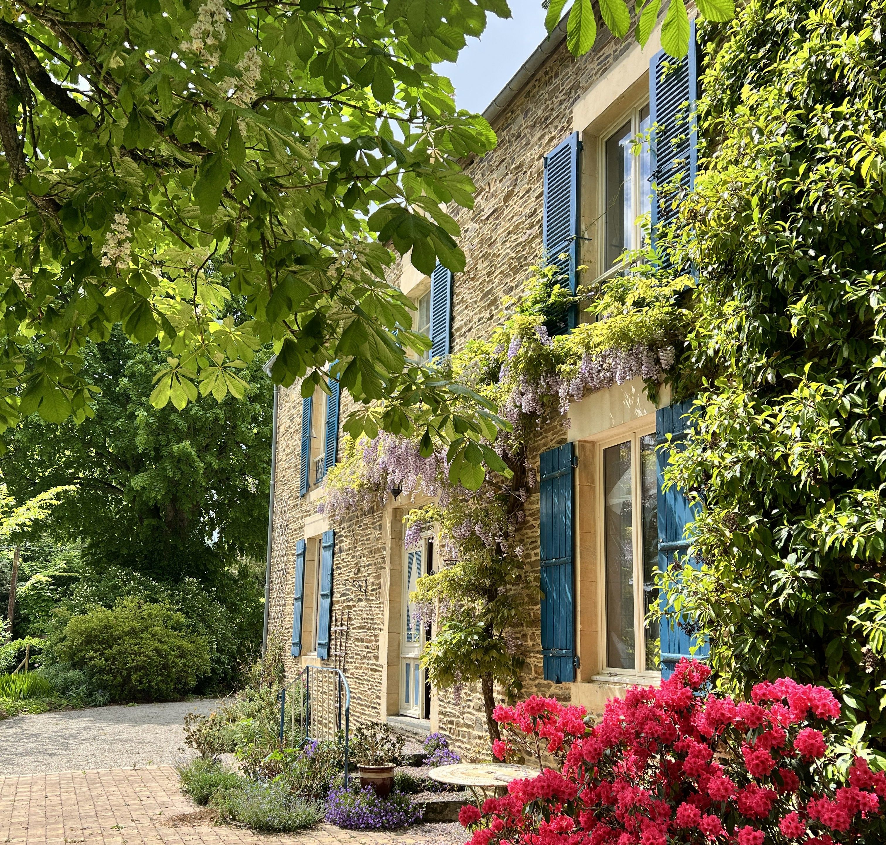 A stone building with blue shutters, surrounded by lush greenery and flowering plants.