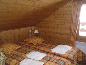 Ein kleiner, gemütlicher Raum mit Holzwänden und einer geneigten Decke. Es gibt ein Bett mit kariertem Überwurf und zwei Kissen, einen Nachttisch mit Lampe sowie ein Fenster mit Vorhängen.