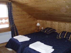 A small, cozy room with a sloped ceiling, wooden walls, a bed with blue bedding, a bedside table with a lamp, and a window with curtains. - Spacious apartment - 8 guests ski-in/ski-out (Les Menuires)