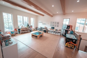 A spacious room with large windows, a play area with toys and a table, and a game table. - Modern 6 Bedroom Northern VA/DC Dream Home! +Gym + Pool + Theater + Playroom (McLean)
