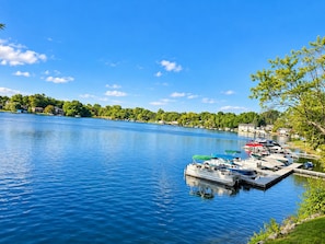 Un llac tranquil amb vaixells amarrats al moll, envoltat de vegetació i cases.