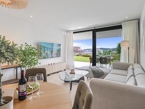 A modern living room with a sofa, a glass coffee table, and a dining area with a table and chairs. There is a TV mounted on the wall, and a view of the outdoors through large windows. - Seaside residence, pets welcome (Estepona)