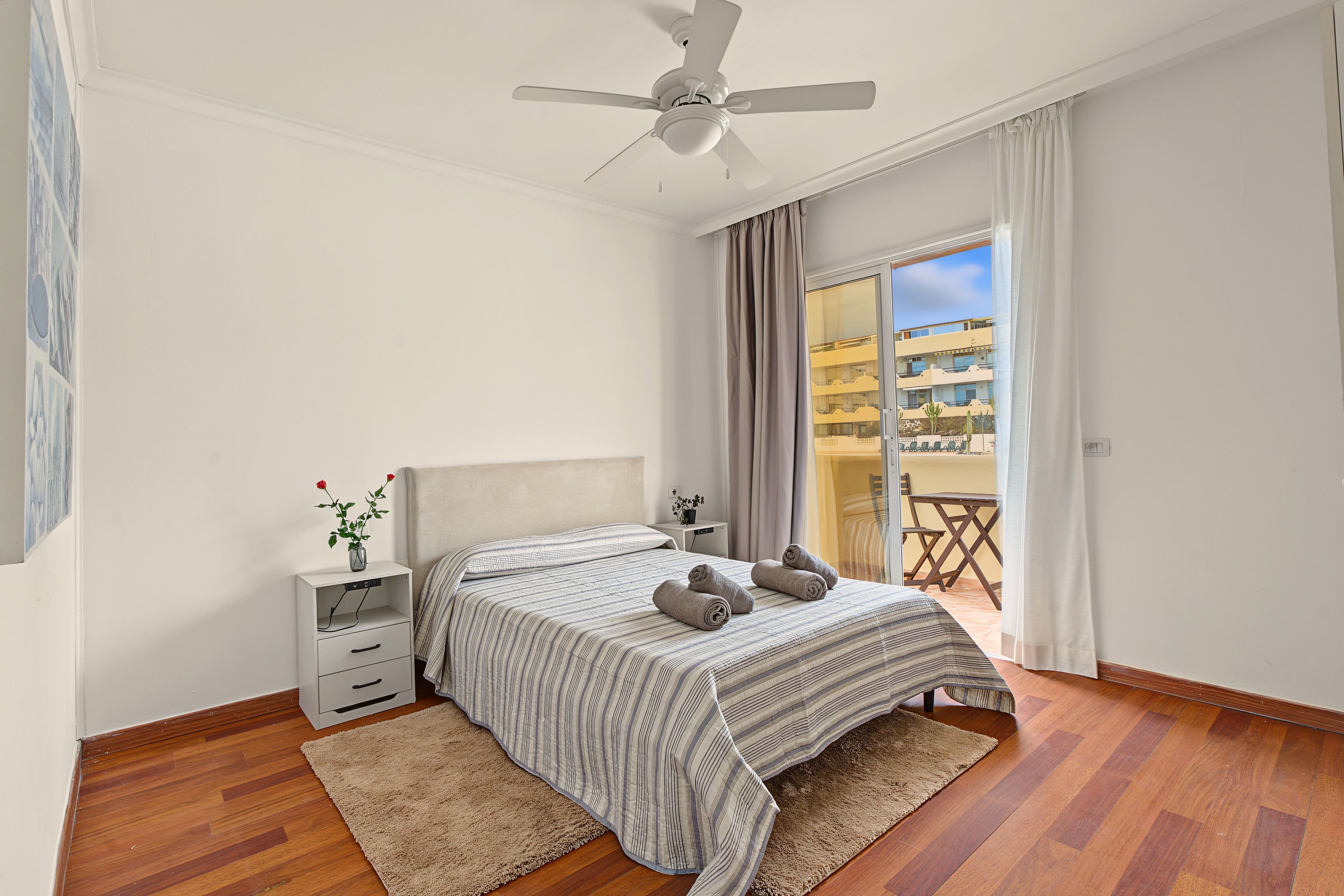 Un dormitorio con cama, mesita de noche, ventilador de techo, ventana con cortinas y vista a un edificio.