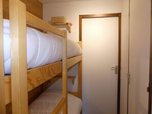 Ein Etagenbett mit Holzrahmen und weißer Bettwäsche, ein kleines Regal mit einem gewebten Gegenstand und eine Tür mit silberner Griffleiste.