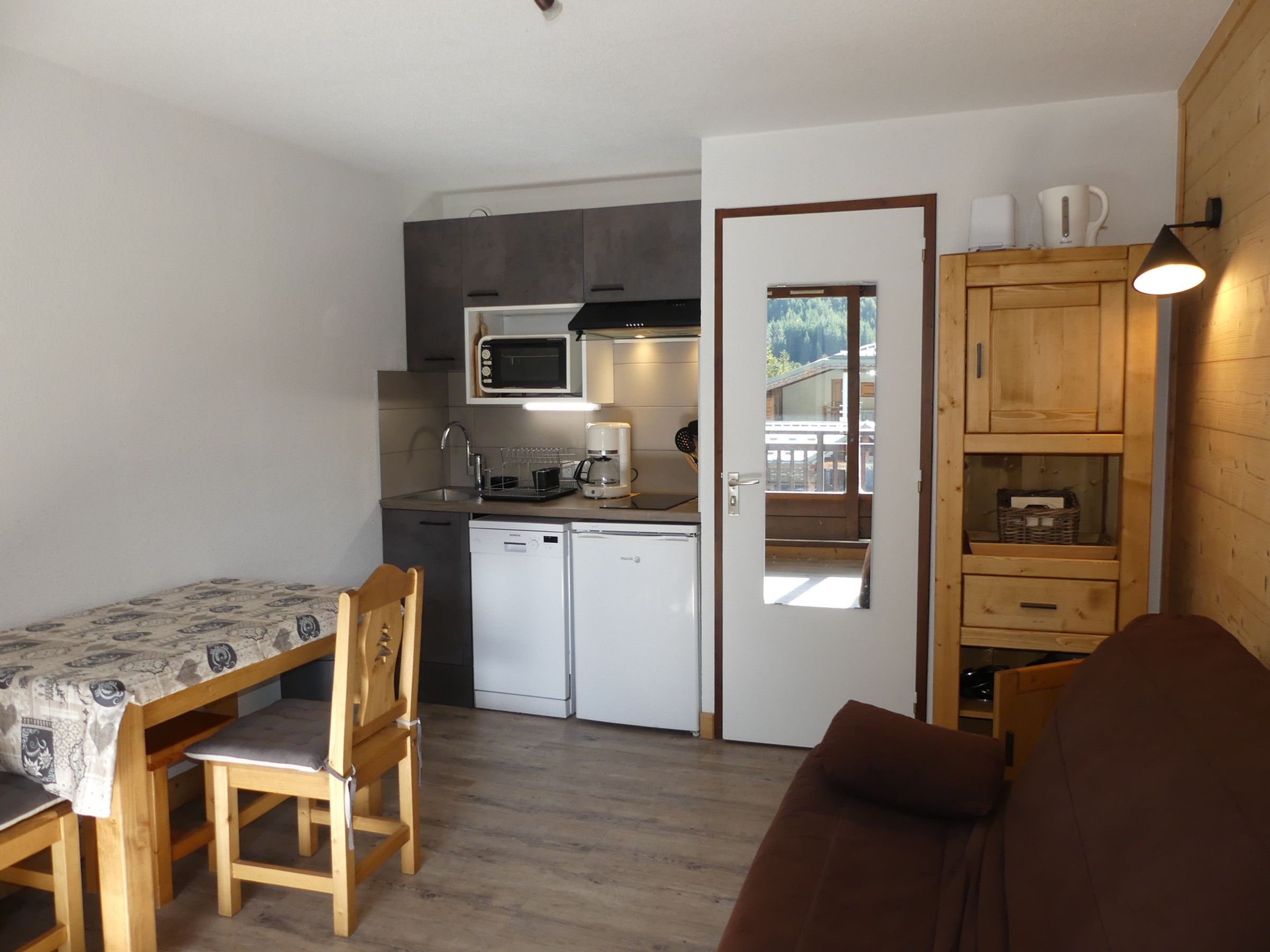 Eine kompakte Küche mit Mikrowelle, Kaffeemaschine und kleinem Kühlschrank. Ein Holztisch mit Stühlen und ein braunes Sofa stehen bereit. Eine Tür führt zu einem Außenbereich mit einer Holzterrasse.