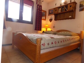 A wooden bed with a quilt, a window with curtains, a wall-mounted shelf with items, and a wall clock. - Chalet in city center - 8 guests (Pralognan-la-Vanoise)