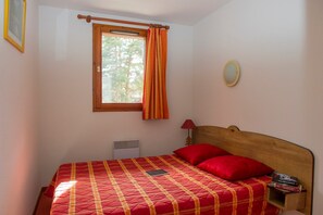 Ein Schlafzimmer mit einem Bett, einem Fenster mit Vorhängen, einem Nachttisch mit Büchern und einem an der Wand befestigten Spiegel.