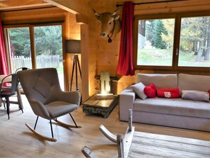 A cozy cabin interior with a rocking chair, a sofa with red cushions, a wooden ceiling, and a large window with a view of trees. - Spacious chalet with terrace - 10 guests (Pralognan-la-Vanoise)