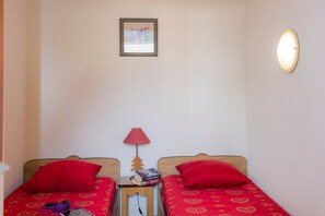 Quarto com duas camas, cada uma com colcha e almofadas vermelhas, uma mesinha com livros, um quadro na parede e um ponto de luz fixado na parede.