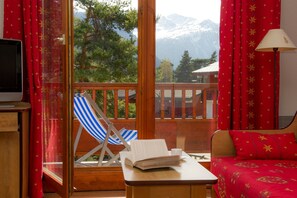 Ein Zimmer mit Holzbalkon, einem blau gestreiften Liegestuhl, einem Bett mit roter Bettwäsche und einem Fernseher auf einer Holzkassette.