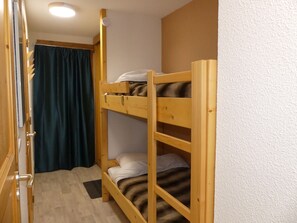 Ein Etagenbett mit Holzrahmen, einer weißen Matratze und einer Streifen-Decke. Eine Tür mit halb zurückgezogenem Vorhang und eine Leuchte an der Decke.