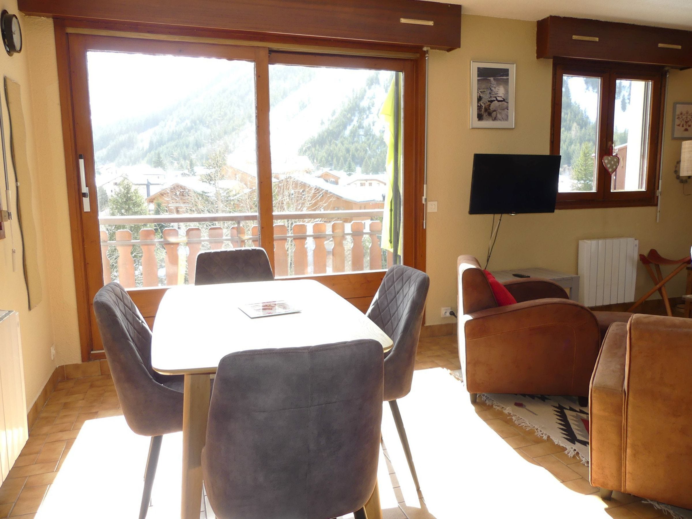 A dining area with a table and chairs, a sofa, a television, and a view of a mountainous landscape through large windows.