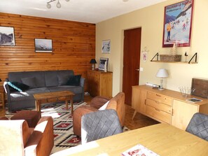 Ein gemütliches Wohnzimmer mit einer Holzwand, einem grauen Sofa, einem Holztisch und einem Esstisch mit Stühlen.