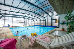An indoor swimming pool with a glass ceiling, surrounded by lounge chairs and a wooden deck. - Ski aux pieds | Proche du télésiège | Piscine (Saint-François-Longchamp)