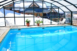 A swimming pool with a glass enclosure, potted plants, and a view of snow-covered buildings. - Au pied des pistes et du télésiège du Marquis (Saint-François-Longchamp)