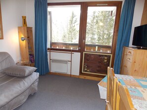 A room with a sofa, a table with a patterned tablecloth, a television on a wooden cabinet, and a window with a view of trees. - Ideal south-facing studio - 4 guests (Pralognan-la-Vanoise)