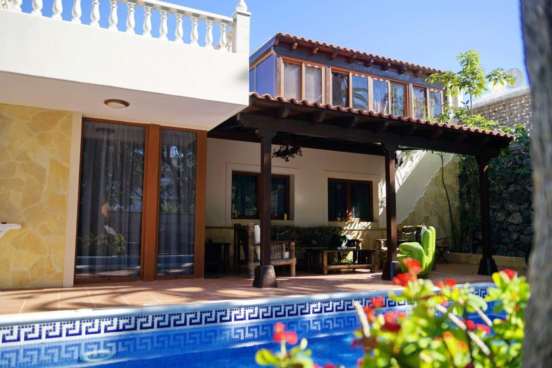 A pool with a geometric tile pattern, a covered patio area with a table and chairs, and a building with a balcony and large windows.