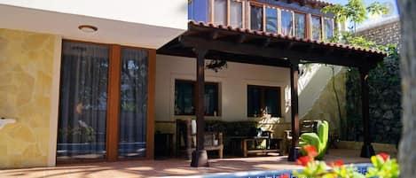 A pool with a geometric tile pattern, a covered patio area with a table and chairs, and a building with a balcony and large windows.