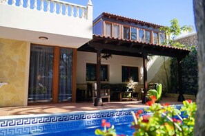 A pool with a geometric tile pattern, a covered patio area with a table and chairs, and a building with a balcony and large windows.