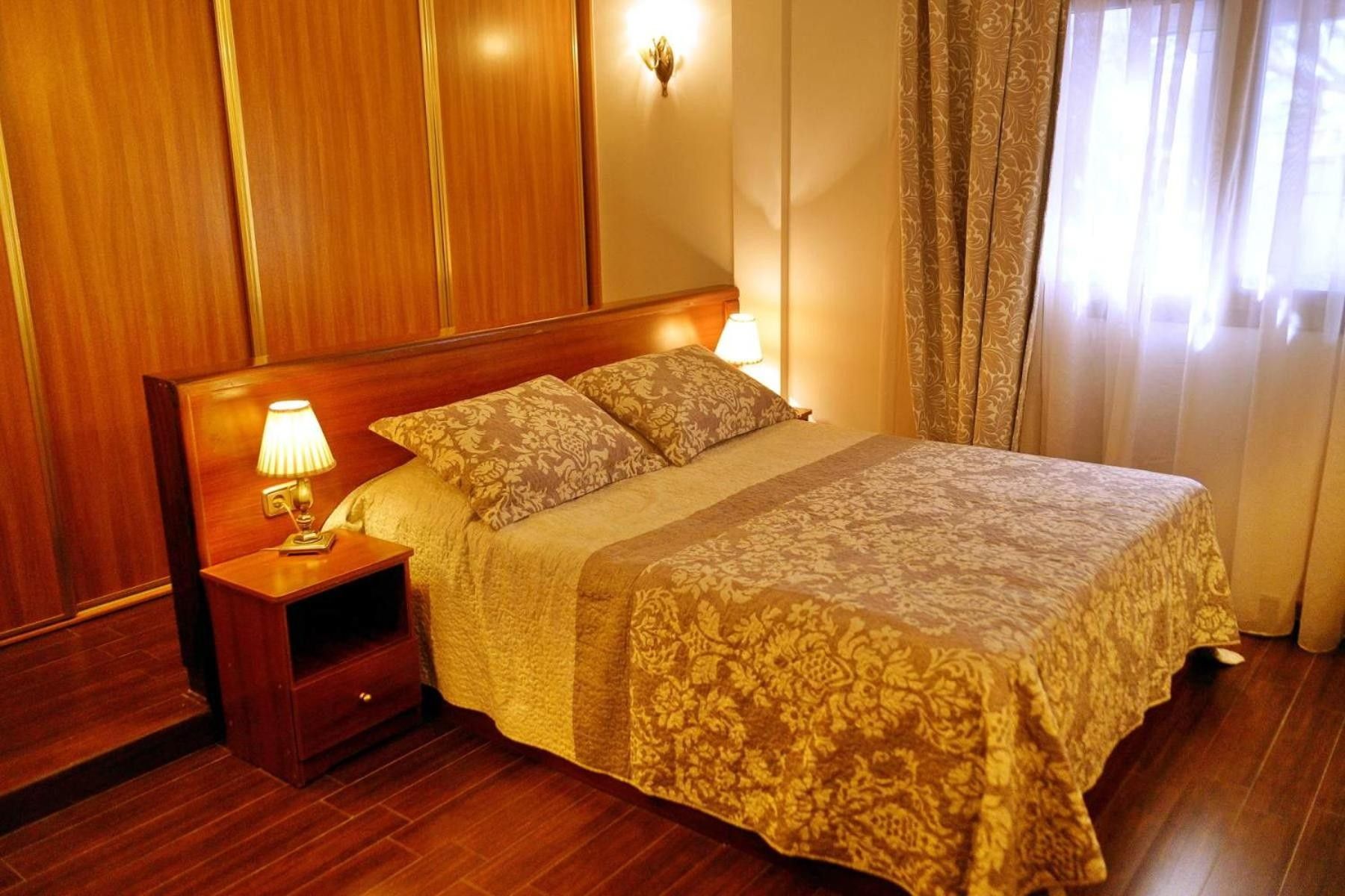 A hotel room with a wooden headboard, a bedside table with a lamp, a bed with patterned bedding, and a window with curtains.