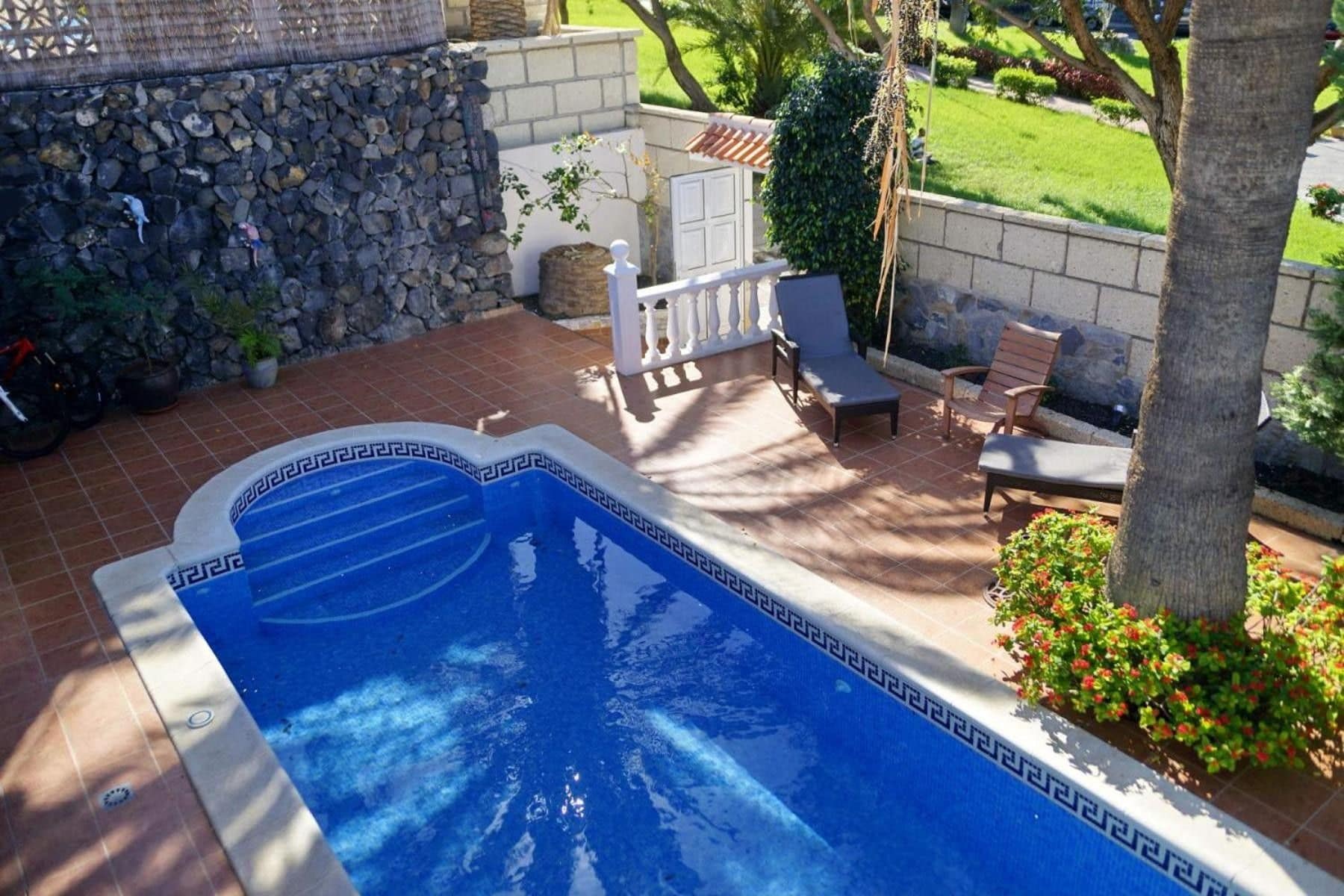 A swimming pool with a stone wall and a patio area with chairs and a table.