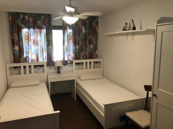 A bedroom with two single beds, a ceiling fan, a nightstand with a lamp, and a wardrobe.