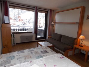 A hotel room with a bed, a sofa, a small table, a lamp, a window with a view of buildings and a balcony, and a radiator. - Studio in the heart of Pralognan - 4 guests (Pralognan-la-Vanoise)