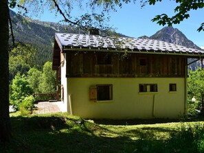 A two-story wooden house with a sloped roof, surrounded by greenery and mountains in the background. - Family chalet with parking - 14 guests (Pralognan-la-Vanoise)