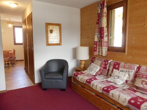 A room with a wooden wall, a sofa with patterned cushions, a chair, a table with a lamp, and a door leading to another room. - Cozy duplex near slopes - 6 guests (Pralognan-la-Vanoise)