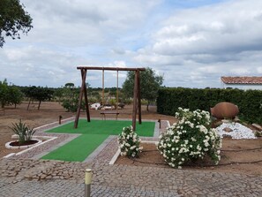 A garden with a wooden swing set, green artificial turf, white flowering bushes, and a paved walkway.