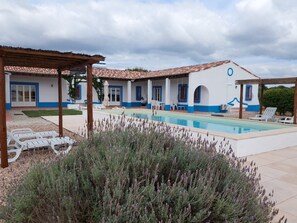 Ein Poolbereich mit einer Pergola, Sitzgelegenheiten im Freien und einem Gebäude mit blauen Verzierungen und weißen Wänden.