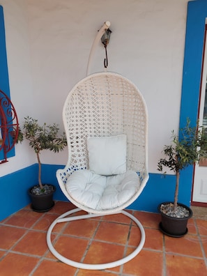 A white wicker hanging chair with a cushion, positioned on a tiled floor with a hose coiled beside it.