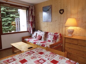 A cozy room with a wooden bed, a wooden table, a sofa with patterned cushions, a lamp, and a poster on the wall. - Cozy apartment with terrace - 5 guests (Pralognan-la-Vanoise)
