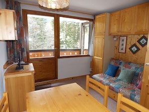 Ein gemütliches Blockhauss-Interieur mit einem Holzespeisetisch, einer Couch mit bunter Stegdecke und einem Fenster mit Blick auf Bäume.