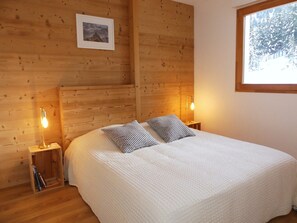 Un dormitorio de cabaña de madera con una cama, dos lámparas de noche, un cuadro en la pared y una ventana con vistas nevadas.