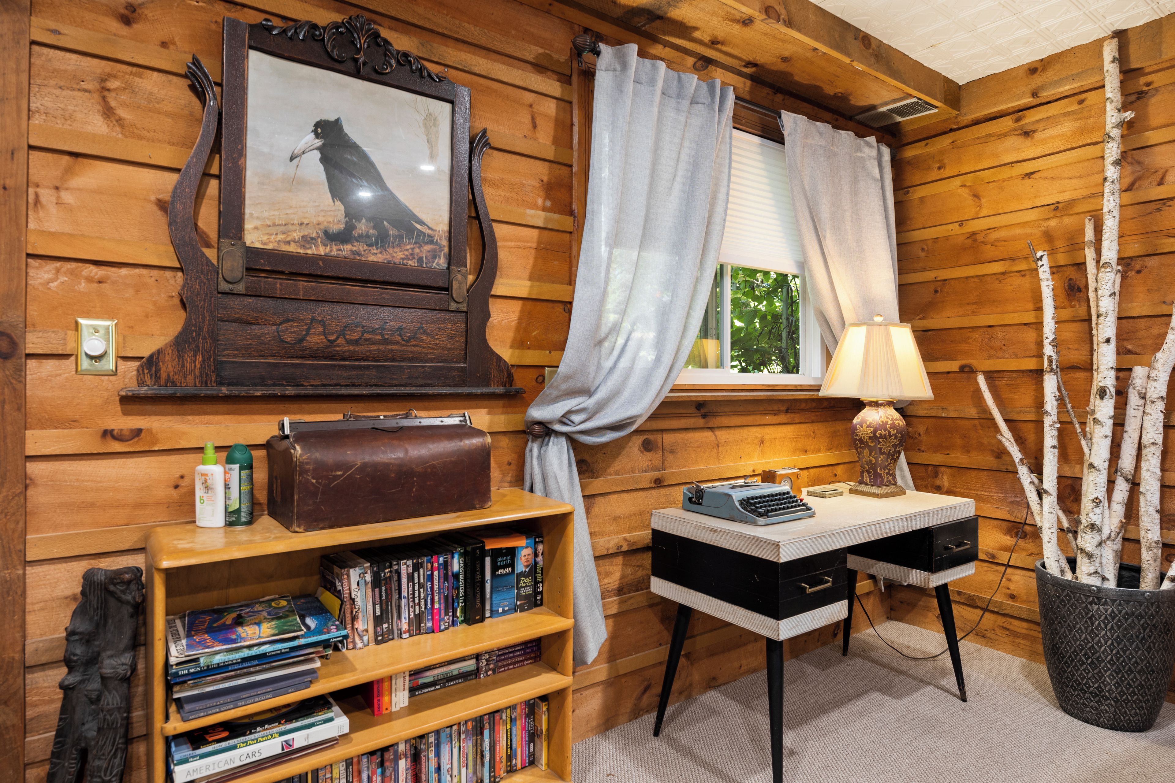 Un interior de madera con una estantería, un escritorio con una lámpara, un cuadro enmarcado y una ventana con cortinas.